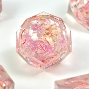 May include: A clear, multi-sided die with pink floral inclusions and glitter. The die has red numbering and is set against a white background. The die is part of a set of dice for tabletop games.