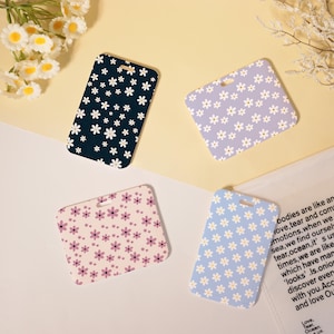 May include: Four rectangular luggage tags with floral patterns. One is navy blue with white daisies, another is lilac with white daisies, a third is pale pink with purple flowers, and the last is light blue with white daisies.
