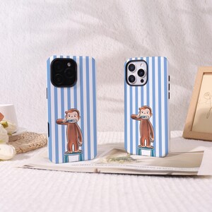 May include: Two smartphone cases featuring a light blue and white vertical striped pattern. Each case displays a cartoon illustration of a brown monkey standing on a small blue stool while brushing its teeth with a blue toothbrush.