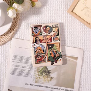 May include: A beige phone case with a comic strip design featuring multiple characters. The case is placed on a newspaper with text, alongside a white floral arrangement and a wooden frame.