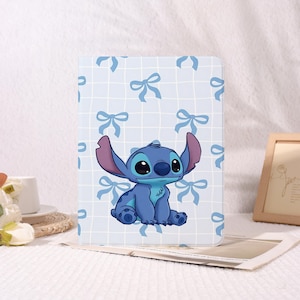 May include: A blue and white tablet case featuring a cartoon illustration of Stitch from Lilo & Stitch. The case has a light blue background with a grid pattern and blue bow accents. Stitch is depicted in a seated position with large ears.