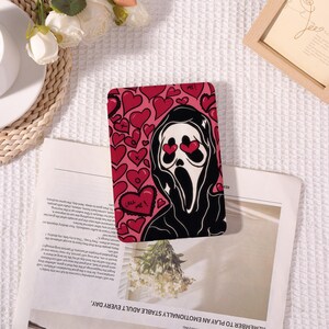 Può includere: Custodia per telefono con un design a volto di fantasma in bianco e nero con occhi a forma di cuore, su sfondo rosa con cuori rossi. Il testo "Call Me!" è visibile. La custodia è appoggiata su un giornale.