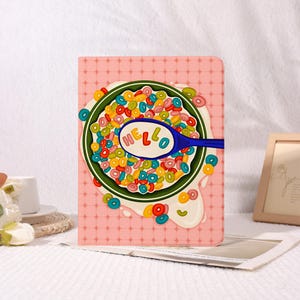 May include: A rectangular tablet case with a pink grid background featuring a graphic of a green bowl filled with colorful ring-shaped cereal. A blue spoon rests in the bowl with the word HELLO printed in colorful letters on the spoon bowl.