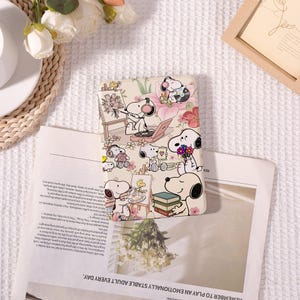 Op de afbeelding: Een rechthoekige beschermhoes voor een e-reader of tablet met een crèmekleurige achtergrond en een herhalend patroon van de hond Snoopy. De illustraties tonen Snoopy die op een doek schildert, naar muziek luistert en bloemen vasthoudt.