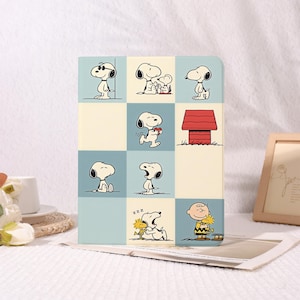 Może przedstawiać: Prostokątne etui na tablet z siatką ilustracji z komiksu Peanuts. Wzór przedstawia Snoopy'ego w różnych pozach, czerwoną budę oraz Charliego Browna z Woodstockiem na tle kwadratów w kolorze kremowym i przygaszonym odcieniu niebieskiego.