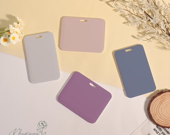 May include: Four rectangular ID badge holders in pastel colors: light gray, mauve, purple, and blue. Each has a small slot at the top. The holders are arranged on a light yellow surface with floral accents and a wooden slice.