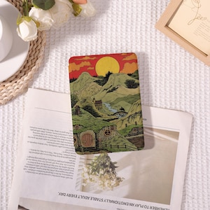 Può includere: Una carta rettangolare con un'illustrazione di paesaggio con dolci colline verdi, un sole al tramonto e case in stile hobbit. La tavolozza dei colori comprende tonalità arancioni, gialle e verdi. La carta è appoggiata su un giornale.