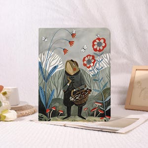 Op de afbeelding: Een decoratief notitieboek met een illustratie van een kikker in een bos. De kikker, gekleed in een grijze jas, draagt een mand. Rode en witte bloemen, paddenstoelen en vlinders zijn verspreid over de scène. De achtergrond is een mix van grijs en groen.