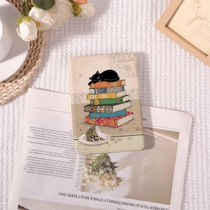 May include: A decorative phone case featuring a stack of colorful books with a black cat resting on top. The case also includes an illustration of a teacup and saucer. The background has a library theme.
