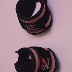May include: Two black ankle weights with pink and white camouflage pattern and adjustable straps. The weights have a black logo that says "The Fitness" with a red wing design.