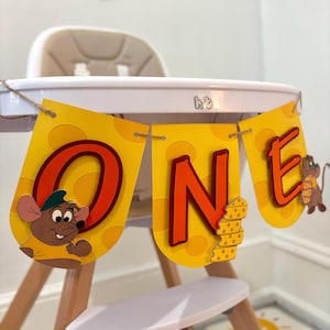 May include: A yellow and orange banner with the word "ONE" in large, red letters. The banner features a cartoon mouse with a green hat and a yellow shirt. The banner is decorated with a cheese pattern.