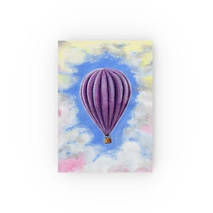 May include: A journal with a painting of a purple hot air balloon against a blue sky filled with fluffy white clouds. The balloon has a detailed, textured design. The background includes soft pastel colours.