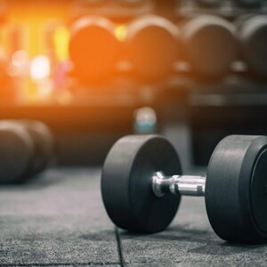 May include: A black dumbbell with a metallic silver handle rests on a dark gray floor. The dumbbell features two circular weights on either side of the central grip. Additional blurred dumbbells are visible in the background.