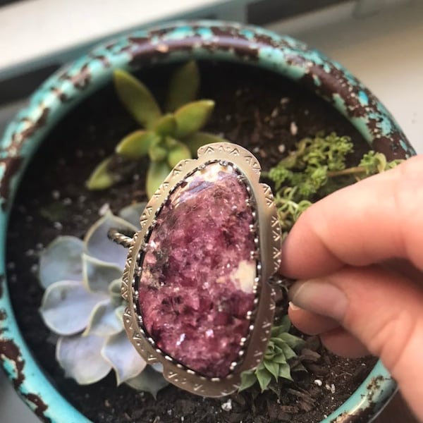 May include: A silver ring with a pink and white gemstone set in a decorative silver bezel. The ring is surrounded by a silver band with a geometric pattern.