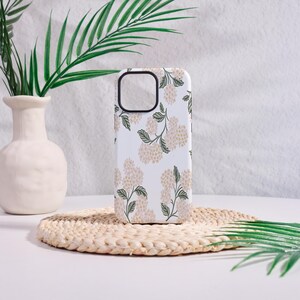 May include: White phone case with a floral pattern featuring beige flower clusters and green leaves. The case has a black camera cutout and is placed on a woven mat, with a white vase and greenery in the background.