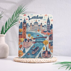 May include: A notebook with a colorful illustration of London landmarks, including Big Ben, St. Paul's Cathedral, and the London Eye. The word "London" is written in a whimsical font across the top. The notebook is resting on a woven coaster.