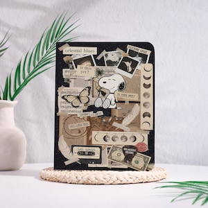Op de afbeelding: Een zwart notitieboek met een collage-ontwerp met Snoopy, een vlinder en diverse vintage elementen. Het notitieboek bevat tekst zoals "celestial blues", "is this yet?" en "Sad Songs". Het notitieboek wordt gepresenteerd op een geweven mat.