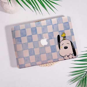 Könnte beinhalten: Eine Laptop-Hülle mit einem hellblauen und beigefarbenen Karomuster. Die Hülle hat eine Cartoon-Illustration von Snoopy und Woodstock. Das Apple-Logo ist sichtbar. Die Hülle liegt auf einer gewebten Matte mit grünen Blättern im Hintergrund.