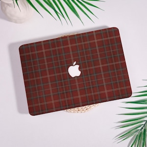 May include: A laptop case with a red plaid pattern, featuring a white Apple logo. The case has a classic tartan design with green and tan lines. The laptop case is on a white surface with decorative plants.