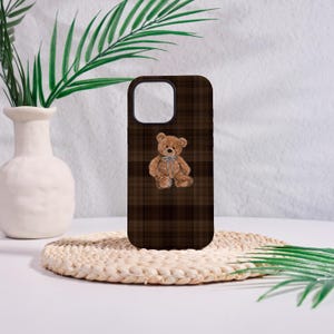May include: A brown phone case featuring a teddy bear illustration with a blue bow. The case has a plaid pattern in shades of brown. The phone case is sitting on a white surface.
