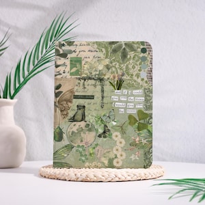 Op de afbeelding: Een decoratief notitieboek met een collage-ontwerp in groen, beige en wit. Het ontwerp bevat vlinders, bloemen en tekstfragmenten. Het notitieboek ligt op een geweven onderzetter.