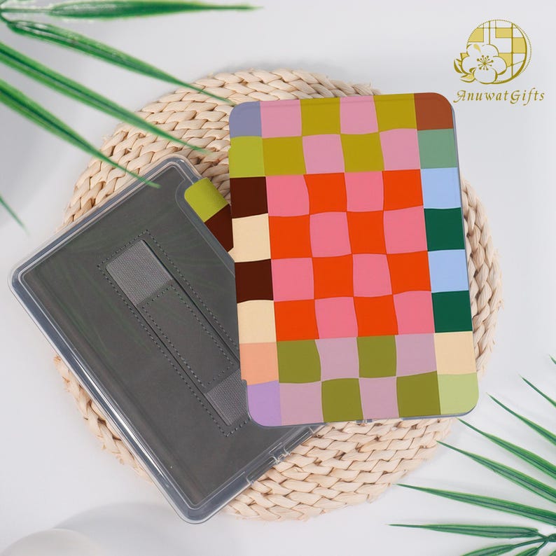 Pode incluir: Uma capa para tablet com um padr&atilde;o xadrez colorido em tons de rosa, laranja, verde e azul. A capa est&aacute; aberta, revelando um interior cinza com uma al&ccedil;a. Descansa sobre uma esteira tecida.