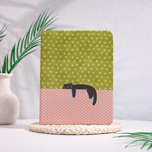 Op de afbeelding: Een rechthoekig notitieboek met een tweekleurig ontwerp op de kaft. Het bovenste gedeelte is olijfgroen met een klein wit bloemenpatroon, terwijl het onderste gedeelte lichtroze is met donkerrode stippen. Een zwarte kat is liggend afgebeeld.