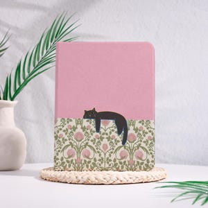 Peut inclure: Un carnet rose avec une illustration de chat noir. La moitié inférieure présente un motif floral crème, rose et vert. Le carnet repose sur un dessous de verre tissé, avec un vase blanc et une plante verte en arrière-plan.