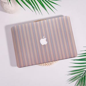 May include: A laptop case with a vertical striped pattern in gray, beige, and red. The Apple logo is visible in the center. The case is on a woven mat, with green palm leaves in the background.