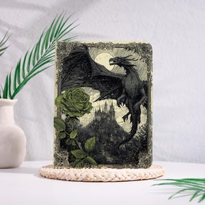 May include: A fantasy-themed journal featuring a black dragon soaring above a castle, with a green rose and moon in the background. The cover has an ornate border and is resting on a woven coaster.