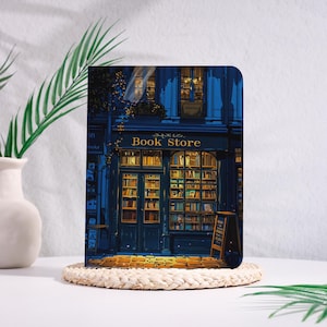 May include: A rectangular print featuring a detailed illustration of a bookstore at night. The building is painted in shades of blue with the words "Book Store" above the entrance. The windows glow with warm light, showcasing shelves of books.