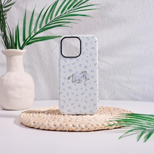 May include: A white phone case with a floral pattern and a cartoon donkey illustration. The case has a black camera cutout and is decorated with small blue flowers. The donkey is gray with a pink bow.