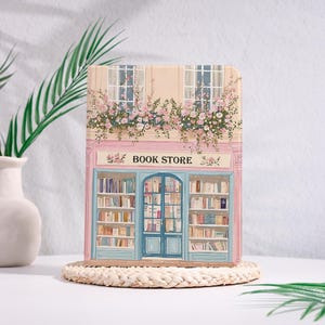May include: A decorative book-shaped item featuring a pastel pink and blue illustration of a bookstore. The facade includes a blue door, windows, and shelves filled with books. The text "BOOK STORE" is above the entrance, with floral accents.