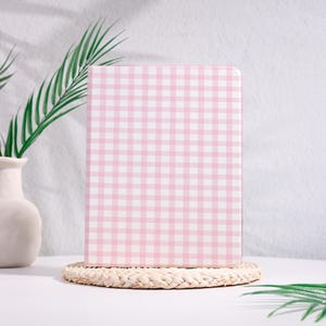 Op de afbeelding: Een roze en wit geruit notitieboek op een geweven onderzetter. Het notitieboek is rechthoekig met afgeronde hoeken. Een witte vaas met groene bladeren staat op de achtergrond.