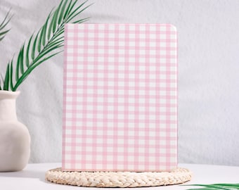 Può includere: Un taccuino rettangolare caratterizzato da un motivo a quadretti vichy rosa chiaro e bianco. Il taccuino è posizionato verticalmente su una base circolare in paglia intrecciata di colore chiaro, con un aspetto pulito e strutturato.