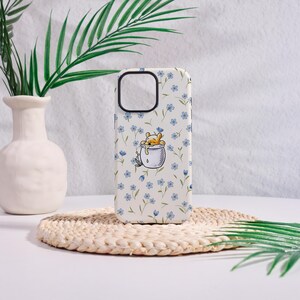 May include: A white phone case featuring a cartoon bear character sleeping in a cup. The case is covered in a pattern of small blue flowers and green stems. The phone case has a black camera cutout.