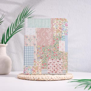 May include: A rectangular notebook with a patchwork design. The cover features various fabric patterns, including floral prints, gingham checks, and solid colors in shades of pink, blue, and green. The notebook is displayed on a woven coaster.