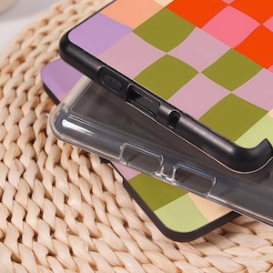 Pode incluir: Duas capas de telem&oacute;vel com um padr&atilde;o xadrez colorido em laranja, rosa, verde e roxo. As capas s&atilde;o pretas e transparentes, com recortes para a c&acirc;mara e bot&otilde;es. As capas est&atilde;o sobre uma superf&iacute;cie tecida.