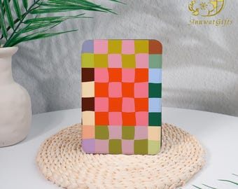 May include: A rectangular coaster with a colorful checkerboard pattern in shades of pink, orange, green, and blue. The coaster is displayed on a woven mat, with a white vase and green plant in the background.