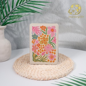 May include: A rectangular, off-white case with a colorful floral design. The flowers are in shades of pink, orange, yellow, and blue, with green leaves. The case is sitting on a woven mat.