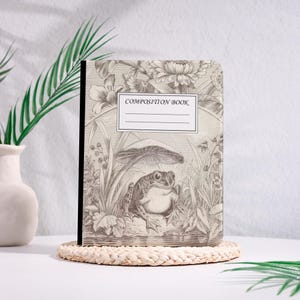 May include: A composition book with a detailed sepia-toned illustration of a frog under a mushroom, surrounded by plants. The cover also features the text "COMPOSITION BOOK" above a blank label area. The book is on a woven coaster.