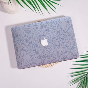 May include: A light purple laptop case with a blue abstract design featuring stars and swirls. The Apple logo is visible. The case is on a woven mat, with green palm leaves in the background.