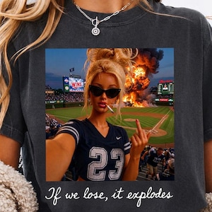 May include: Dark gray t-shirt featuring a graphic of a woman in sunglasses holding a cigarette, with a baseball stadium in the background. The text "If we lose, it explodes" is printed below the graphic.