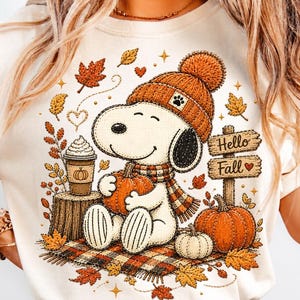 May include: A cream-colored t-shirt featuring a graphic of a cartoon dog wearing an orange knit hat and scarf, holding a pumpkin. The design includes a coffee cup, a wooden sign reading Hello Fall, scattered autumn leaves, and a plaid blanket.