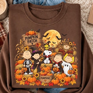 May include: A brown long-sleeve sweatshirt featuring a graphic print of Peanuts characters in Halloween costumes at a pumpkin patch. The design includes a full moon, bats, sunflowers, and the text Pumpkin Patch. The sweatshirt is layered over blue jeans.