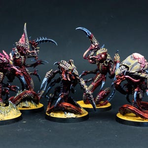 Peut inclure: Cinq figurines de créatures extraterrestres miniatures avec des corps rouges segmentés, des griffes pointues aux extrémités bleues et des carapaces texturées. Chaque figurine repose sur une base noire circulaire avec une surface jaune texturée.