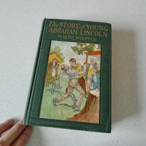 May include: A vintage hardcover book titled "The Story of Young Abraham Lincoln" by Wayne Whipple. The cover is green with a colorful illustration of a young man in a fight. The book is on a white surface.