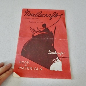 May include: A vintage "Needlecraft" book cover in red with a silhouette of a person doing needlework. The cover also features the text "Embroidery and Fancywork" and "Book of Materials".