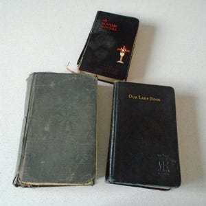 May include: Three vintage books are displayed on a white surface. The books are in various states of wear, with dark covers. One book is titled "My Sunday Missal" with a cross illustration. Another is labeled "Our Lady Book". The third book has a faded, gray cover.