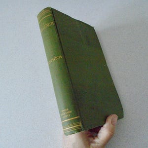 May include: A vintage book with a green cover and gold lettering. The spine reads "Hellenism" and "Nantwich". The book is held at an angle, showcasing its aged appearance and the text on the spine.